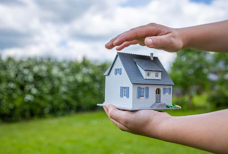 Versicherungen beim Hausbau: Person hält schützend eine Hand über ein Modellhaus